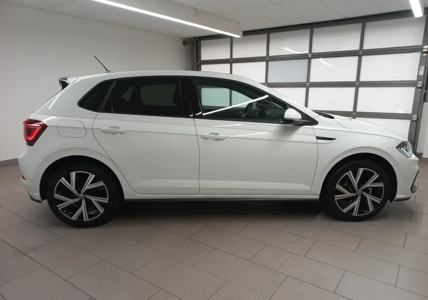 Vue de profil côté gauche d'une Volkswagen Polo blanche avec vitres arrière surteintées et jantes R-Line noires et argentées.