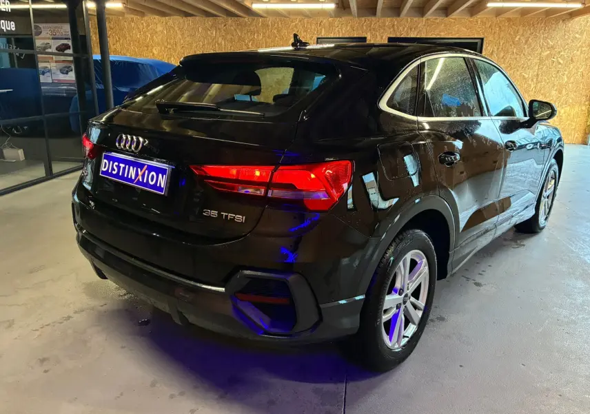 Audi Q3 Sportback noir vu en 3/4 arrière droit, avec feux arrière allumés et jantes alu visibles en intérieur showroom.