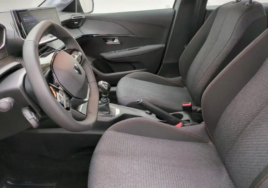 Vue intérieure côté conducteur de la Peugeot 208 2025, sièges tissu gris avec surpiqûres orange et volant cuir compact.