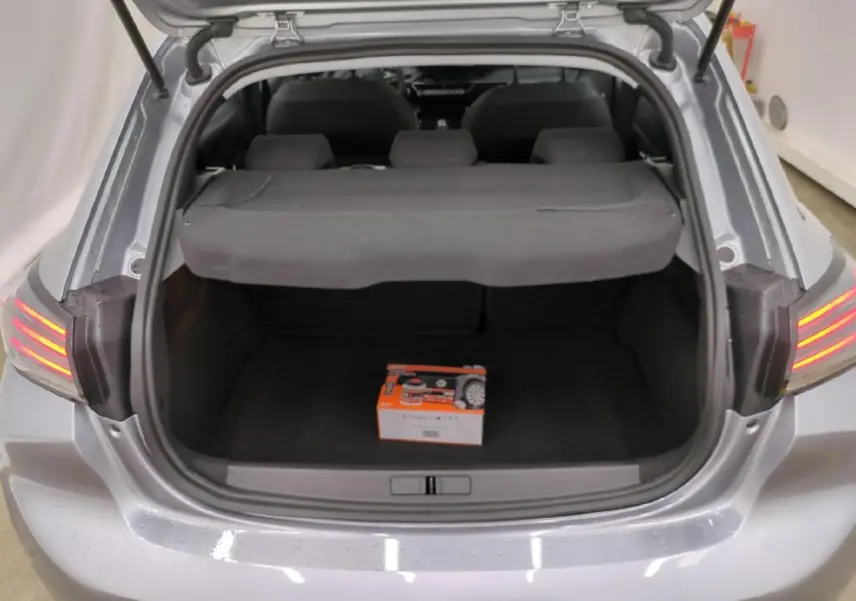Coffre ouvert vu de l'arrière d'une Peugeot 208 gris clair, avec cache-bagages et boîte orange à l'intérieur.
