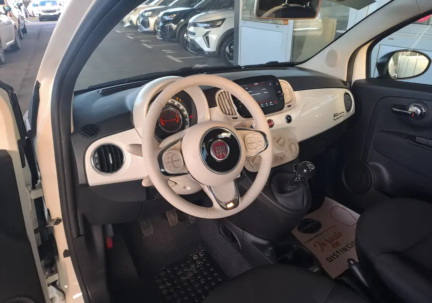 Intérieur côté conducteur d'une Fiat 500 blanche 2023, volant beige, tableau de bord compact avec écran tactile et levier de vitesses manuel.