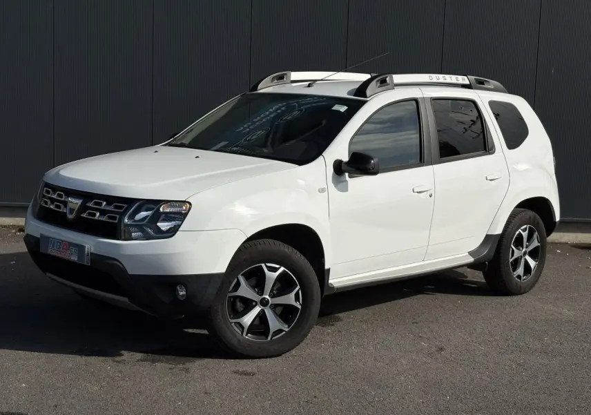 Dacia Duster blanc glacier en 3/4 avant droit avec barres de toit alu et jantes diamantées noires.