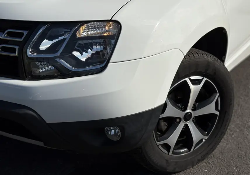 Gros plan sur l'avant droit du Dacia Duster blanc glacier, mettant en valeur le phare et la jante alu diamantée noire.