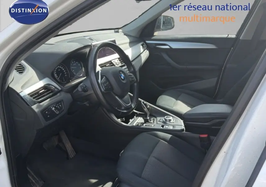 Vue intérieure côté conducteur d'une BMW X1 blanche, mettant en valeur le tableau de bord et les sièges noirs.