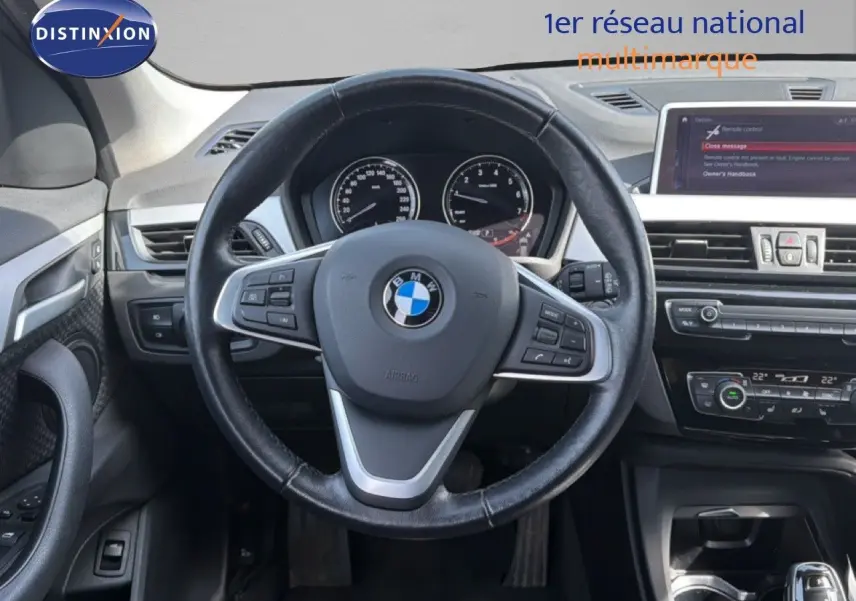 Vue intérieure centrée sur le volant noir d'une BMW X1 blanche, avec tableau de bord et écran multimédia visibles.