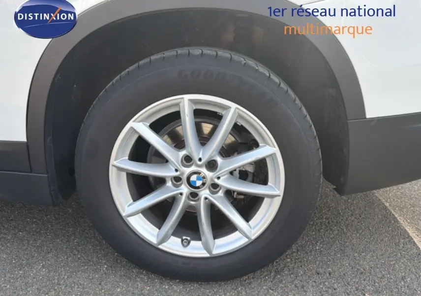 Gros plan sur la roue avant droite d’un BMW X1 blanc, avec jante alliage argentée à dix branches.