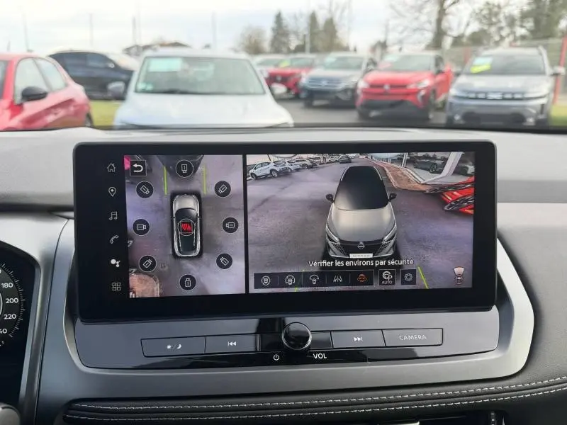 Vue intérieure du tableau de bord du Nissan Qashqai Hybrid e-POWER 2025 montrant l'écran de caméra 360° avec vue aérienne du véhicule blanc.