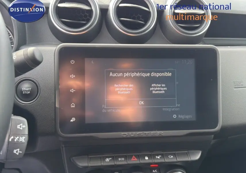 Vue rapprochée de l’écran tactile et de la console centrale du Dacia Duster bleu Iron, avec commandes et aérateurs ronds visibles.