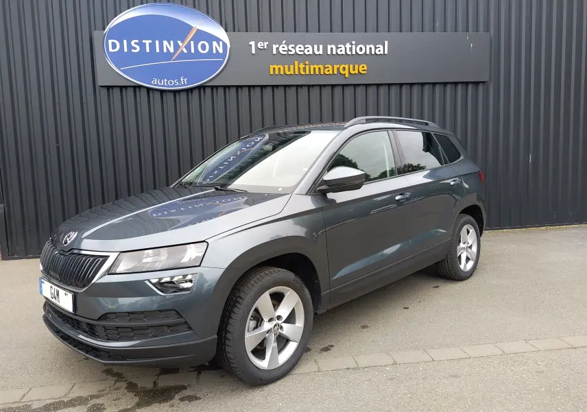 Vue 3/4 avant droite d’un Skoda Karoq gris 2021 avec jantes alliage et toit ouvrant, devant un mur noir avec enseigne Distinxion.