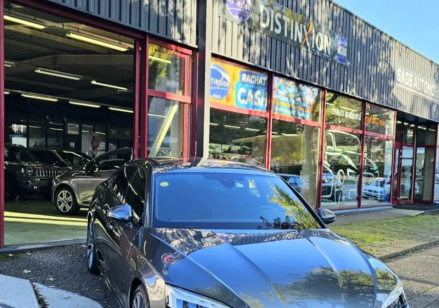 Audi A5 Sportback gris Daytona vue en 3/4 avant droit devant un garage avec enseignes visibles.