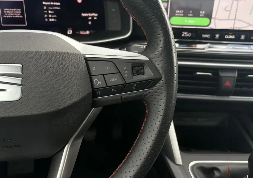 Gros plan sur le volant en cuir noir à surpiqûres rouges du SEAT Leon 2025, avec commandes multifonctions et écran tactile en arrière-plan.