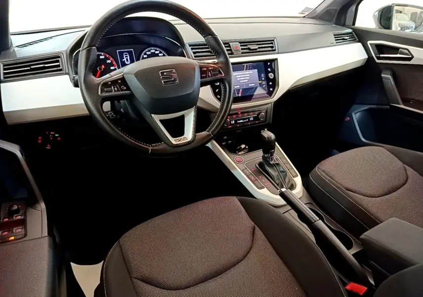 Vue intérieure avant du tableau de bord et volant cuir alu d’un SEAT Arona gris clair avec écran tactile central allumé.