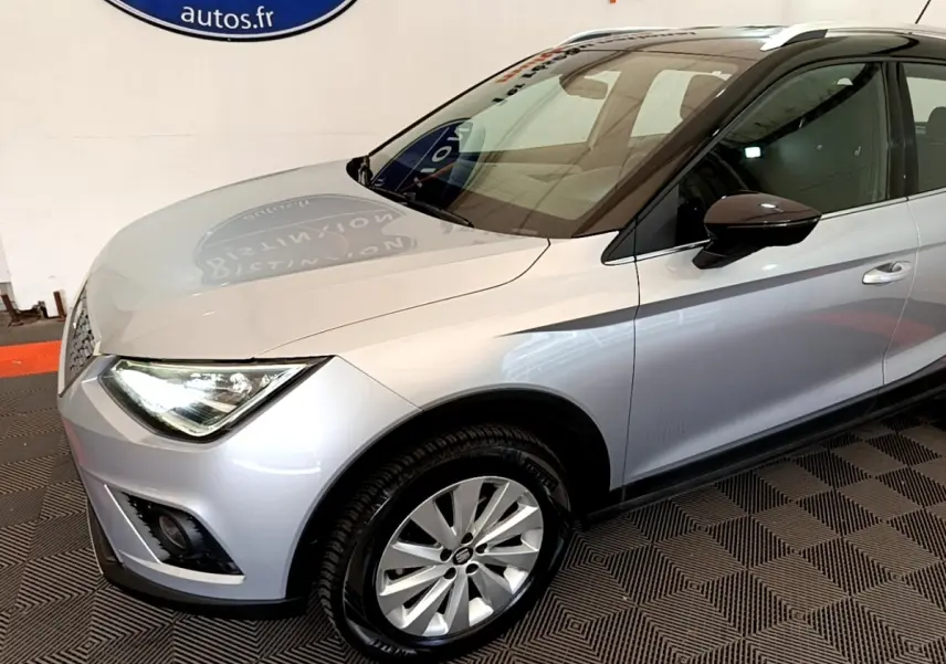 SEAT Arona gris clair vu de 3/4 avant droit, avec phares LED et jantes alliage visibles en intérieur showroom.