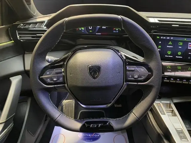 Vue rapprochée du volant noir perforé de la Peugeot 308 GT hybride 2026 avec tableau de bord digital et écran tactile.