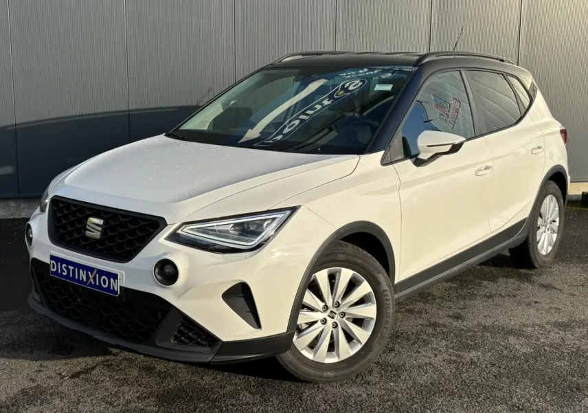 Vue 3/4 avant droite d'un SEAT Arona blanc avec toit noir et jantes alliage 16 pouces, stationné devant un mur gris.