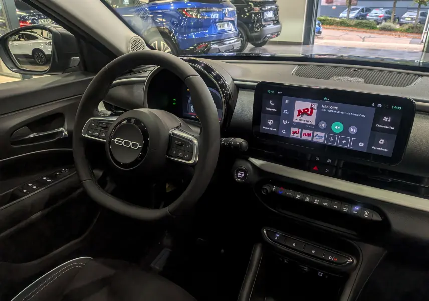 Intérieur de la FIAT 600 Hybrid 2025 vu côté conducteur, volant rond avec logo 600 et écran tactile central allumé.