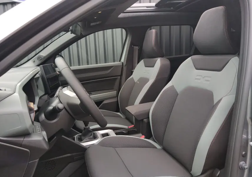 Intérieur du Dacia Bigster 2025 vu côté gauche, sièges bi-ton gris avec logo embossé et toit ouvrant panoramique.