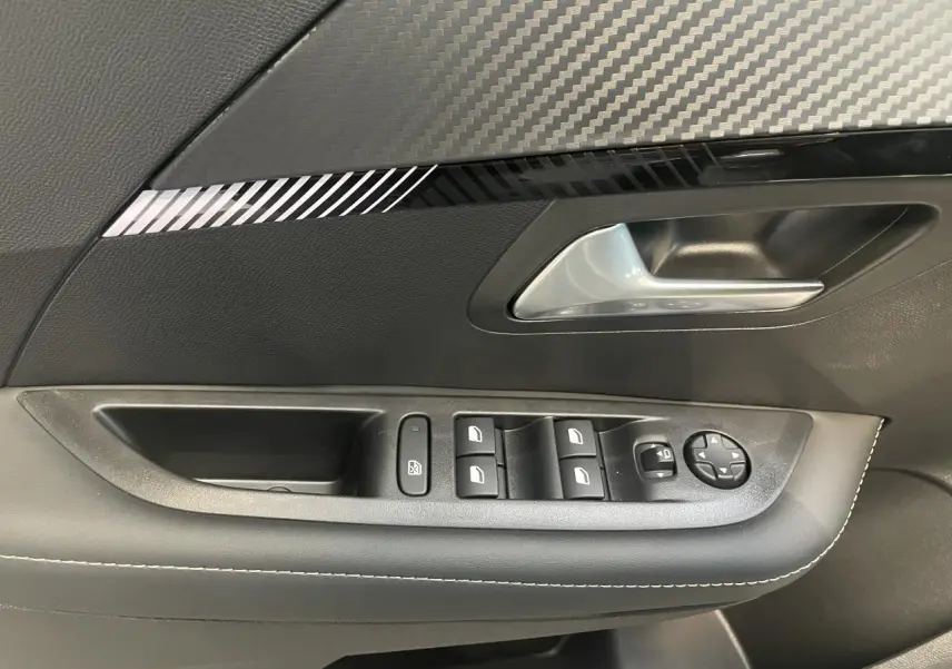Poignée de porte intérieure côté gauche avec commandes de vitres et décor carbone sur Peugeot 208 Gris Artense 2025.
