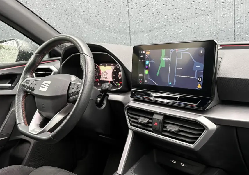 Vue intérieure côté conducteur du tableau de bord du SEAT Leon 2024 avec écran tactile et volant cuir noir à surpiqûres rouges.