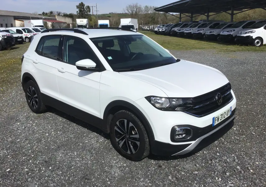 Volkswagen T-Cross blanc vue 3/4 avant droit, avec jantes noires et toit ouvrant, stationné sur un sol gravillonné.
