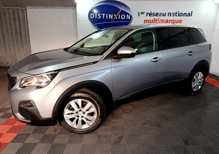 Peugeot 5008 gris clair vue 3/4 avant droit en intérieur, avec jantes alliage et feux allumés.
