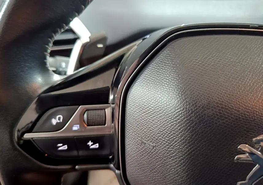 Gros plan sur le volant cuir noir du Peugeot 5008 gris clair, avec commandes multifonctions et logo lion visible.