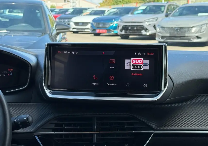 Vue intérieure centrée sur l'écran tactile du tableau de bord de la Peugeot 208 gris Artense, avec vue sur le pare-brise et voitures au loin.