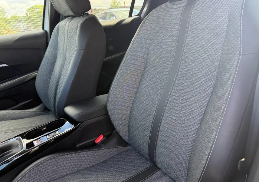 Vue rapprochée des sièges avant en tissu gris avec surpiqûres, accoudoir central et console noire brillante Peugeot 208 2025.