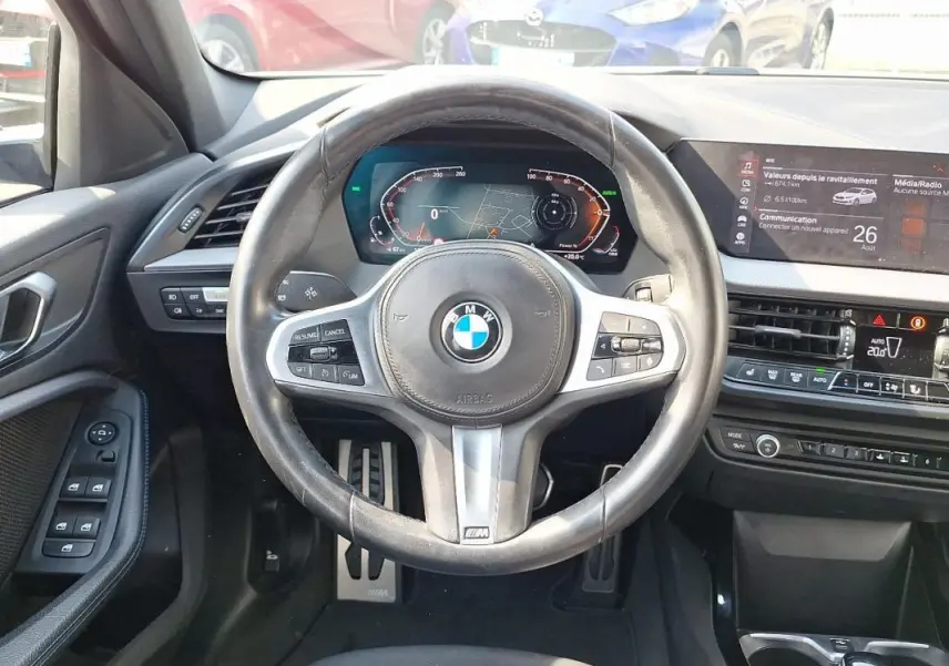 Vue intérieure centrée sur le volant cuir multifonctions et tableau de bord numérique d'une BMW Série 1 blanche 2021.