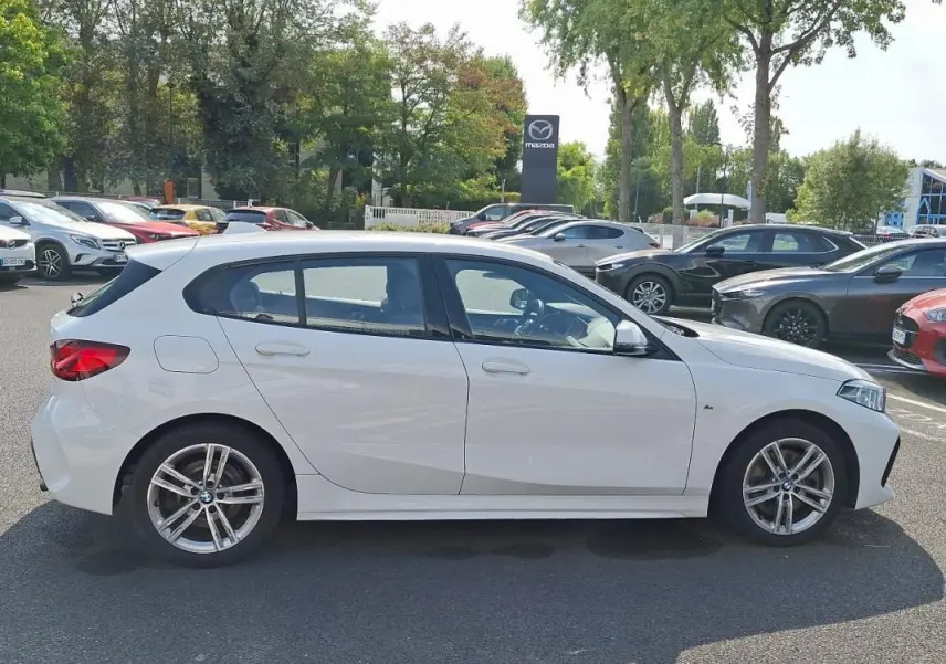 Profil côté gauche d'une BMW Série 1 blanche 120i M Sport 2021, avec jantes aluminium et lignes dynamiques visibles.