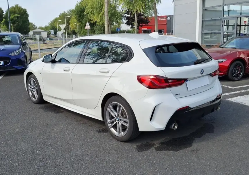 BMW Série 1 120i M Sport blanc vue 3/4 arrière droit, avec jantes aluminium et double sortie d’échappement chromée.