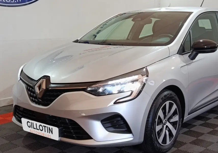 Vue 3/4 avant droite d'une Renault Clio gris clair 2023 avec phares LED allumés et jantes distinctives.