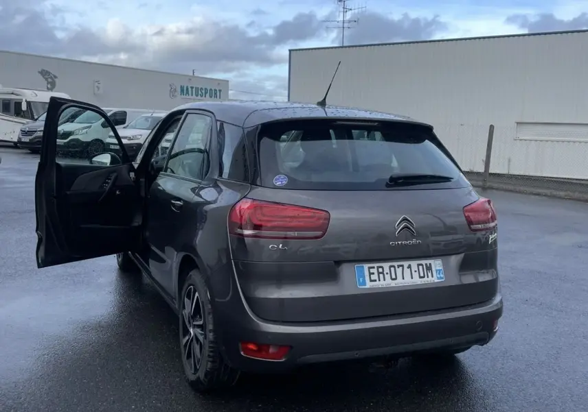 Vue 3/4 arrière gauche d'une Citroën C4 Picasso marron 2017 avec porte avant gauche ouverte sur parking humide.