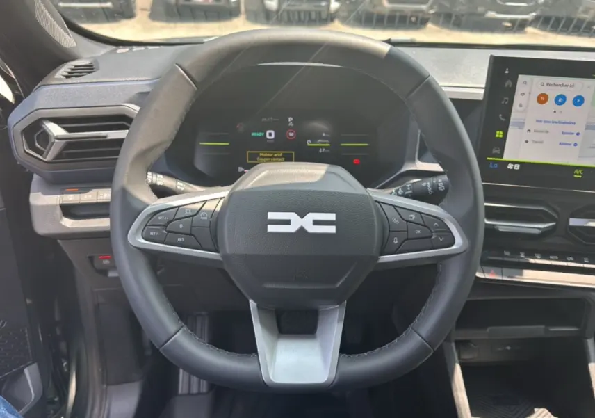 Vue intérieure centrée sur le volant cuir noir du Dacia Duster HEV 140 Journey Plus avec écran tactile et tableau de bord numérique.
