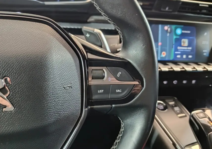 Gros plan sur le volant cuir côté droit de la Peugeot 508 gris clair, avec commandes audio et écran tactile en arrière-plan.