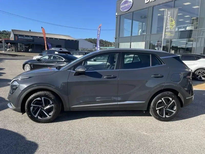 Vue de profil côté gauche d'un KIA Sportage gris Eclipse Métal 2023 avec jantes alliage bicolores sur parking extérieur.