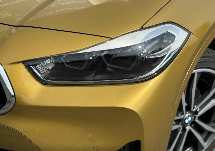 Gros plan sur le phare avant droit et la calandre d'une BMW X2 F39 or Galvanic avec jante alliage bicolore visible.