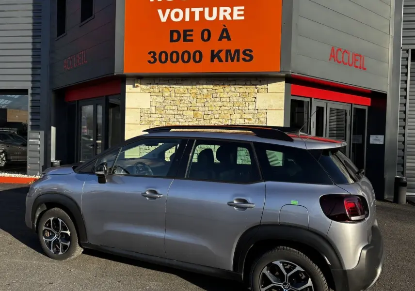 Vue latérale droite d'un Citroën C3 Aircross gris clair garé devant un bâtiment avec enseigne orange "Votre nouvelle voiture".