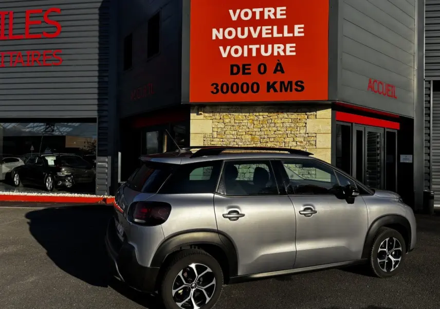 Citroën C3 Aircross gris clair vue de profil côté droit, stationnée devant un bâtiment avec panneau orange.