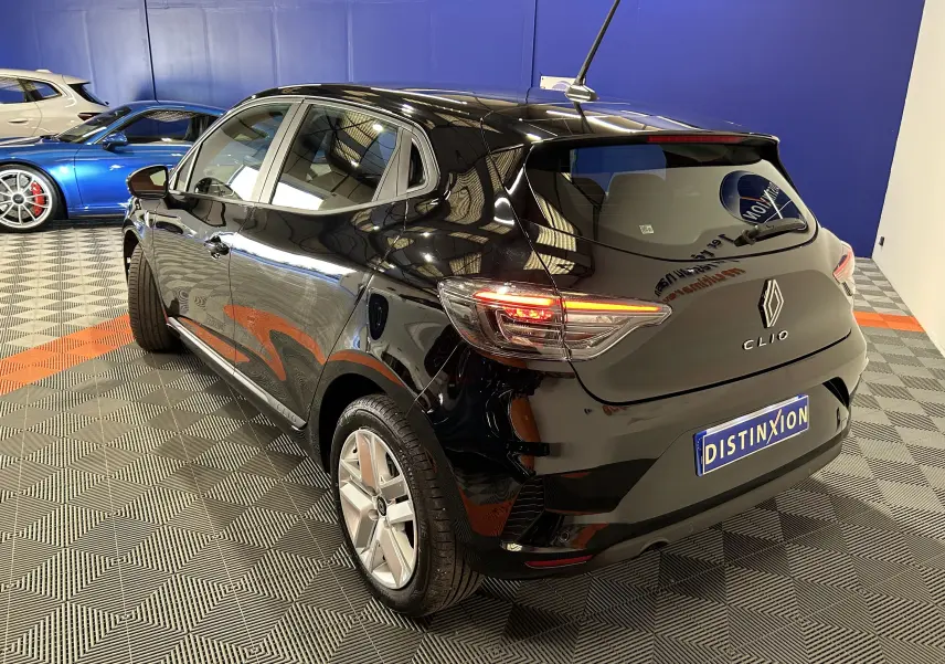 Vue 3/4 arrière droite d'une Renault Clio V noire, avec feux arrière allumés et finition brillante dans un showroom.