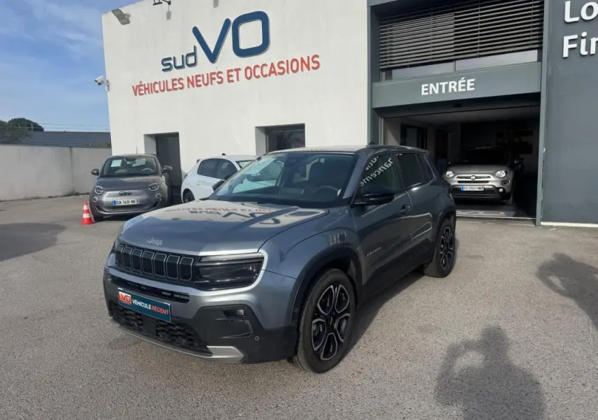 Jeep Avenger gris en 3/4 avant droit, SUV compact avec calandre noire et jantes distinctives, stationné devant un garage.