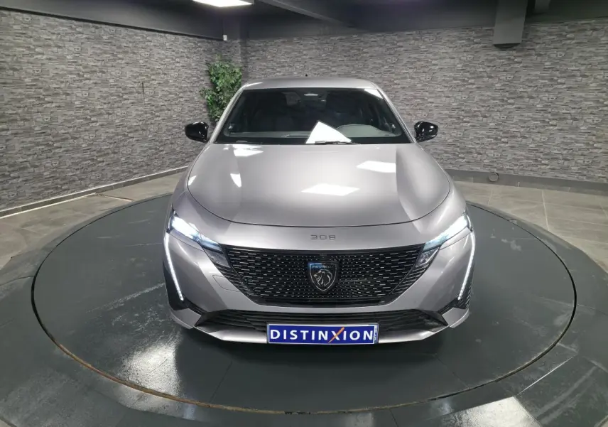 Peugeot 308 gris métallisé vue de face, avec calandre noire brillante et feux LED allumés dans un showroom.