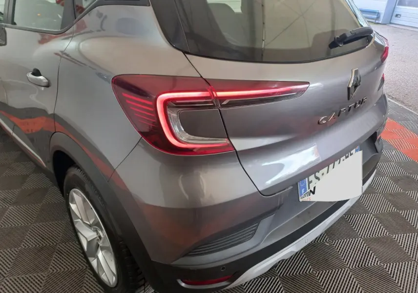 Vue 3/4 arrière droite d'un Renault Captur gris clair avec feux arrière LED et logo visible sur un sol à damiers.