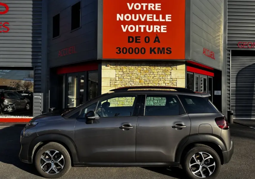 Profil latéral gauche d'un Citroën C3 Aircross gris foncé garé devant un bâtiment avec enseigne rouge.