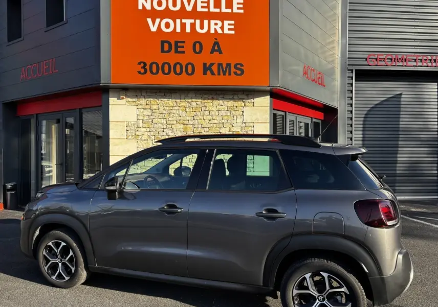 Profil droit d'un Citroën C3 Aircross gris foncé 2024 avec barres de toit noires devant un bâtiment commercial.