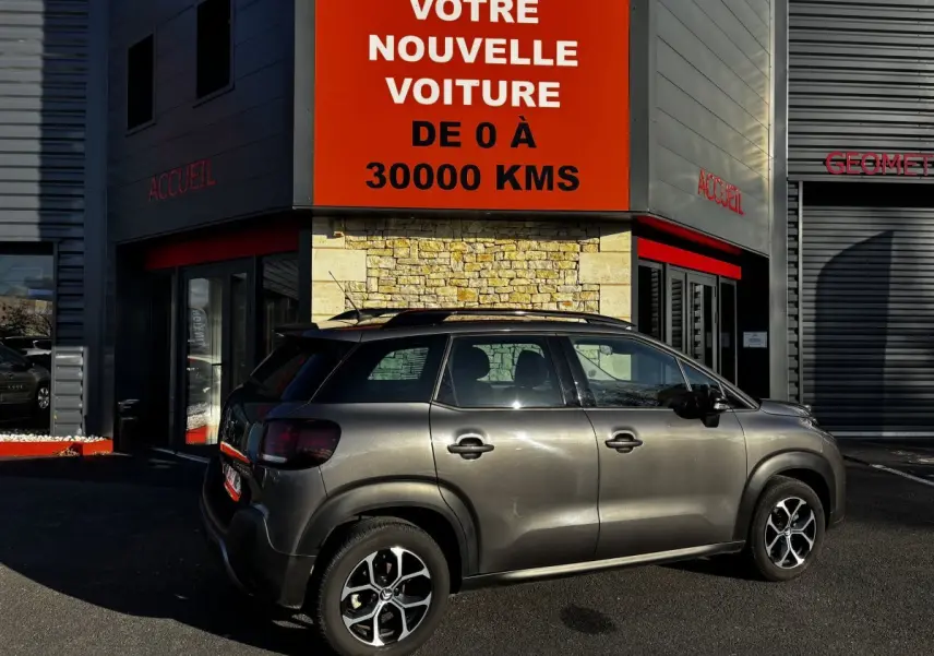 Vue latérale droite d'un Citroën C3 Aircross gris foncé 2024 garé devant un bâtiment avec enseigne rouge.