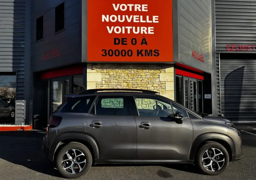 Profil côté droit d'un Citroën C3 Aircross gris foncé 2024 garé devant un bâtiment avec panneau rouge "Votre nouvelle voiture".
