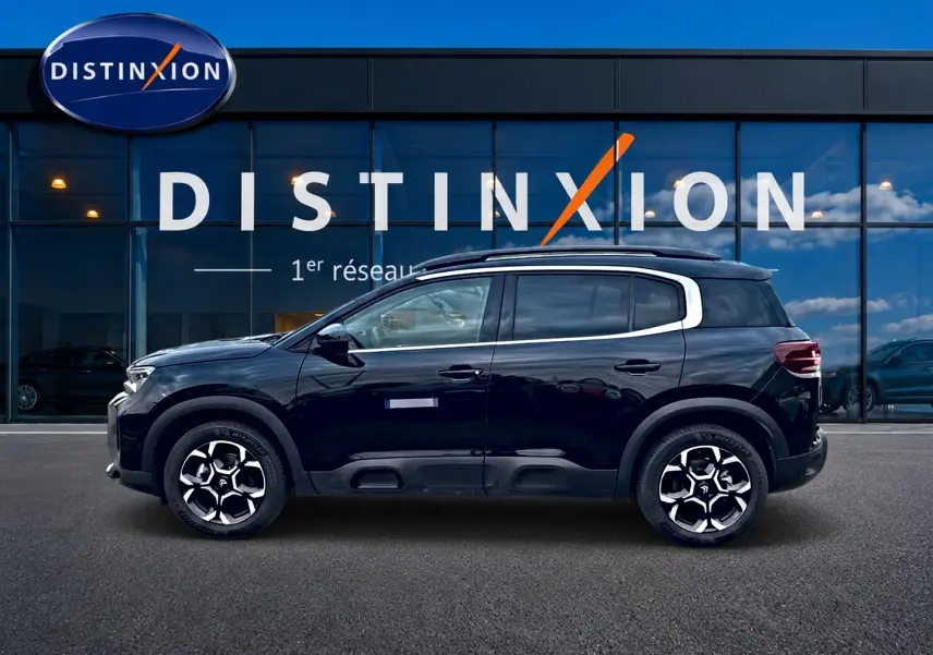Profil côté gauche d'un Citroën C5 Aircross noir 2025 avec jantes alu et barres de toit visibles devant une vitrine Distinxion.