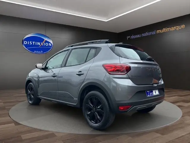 Vue 3/4 arrière gauche d'une Dacia Sandero Stepway TCE 110 Extreme Plus gris urban avec jantes alu noires et barres de toit noires.