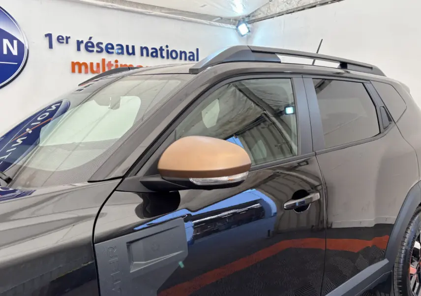 Dacia Duster noir vue côté gauche avant, avec rétroviseur couleur bronze et barres de toit noires.
